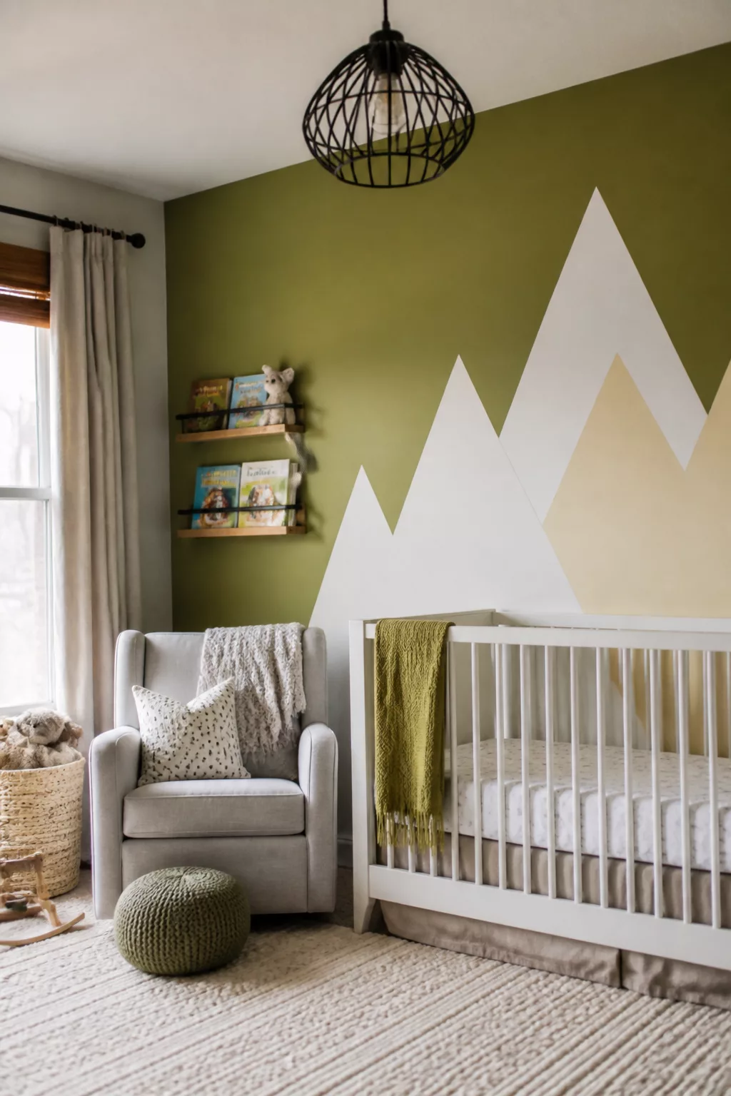Olive green accent wall nursery with geometric painted mountain mural, white crib, gray glider, and knit pouf.