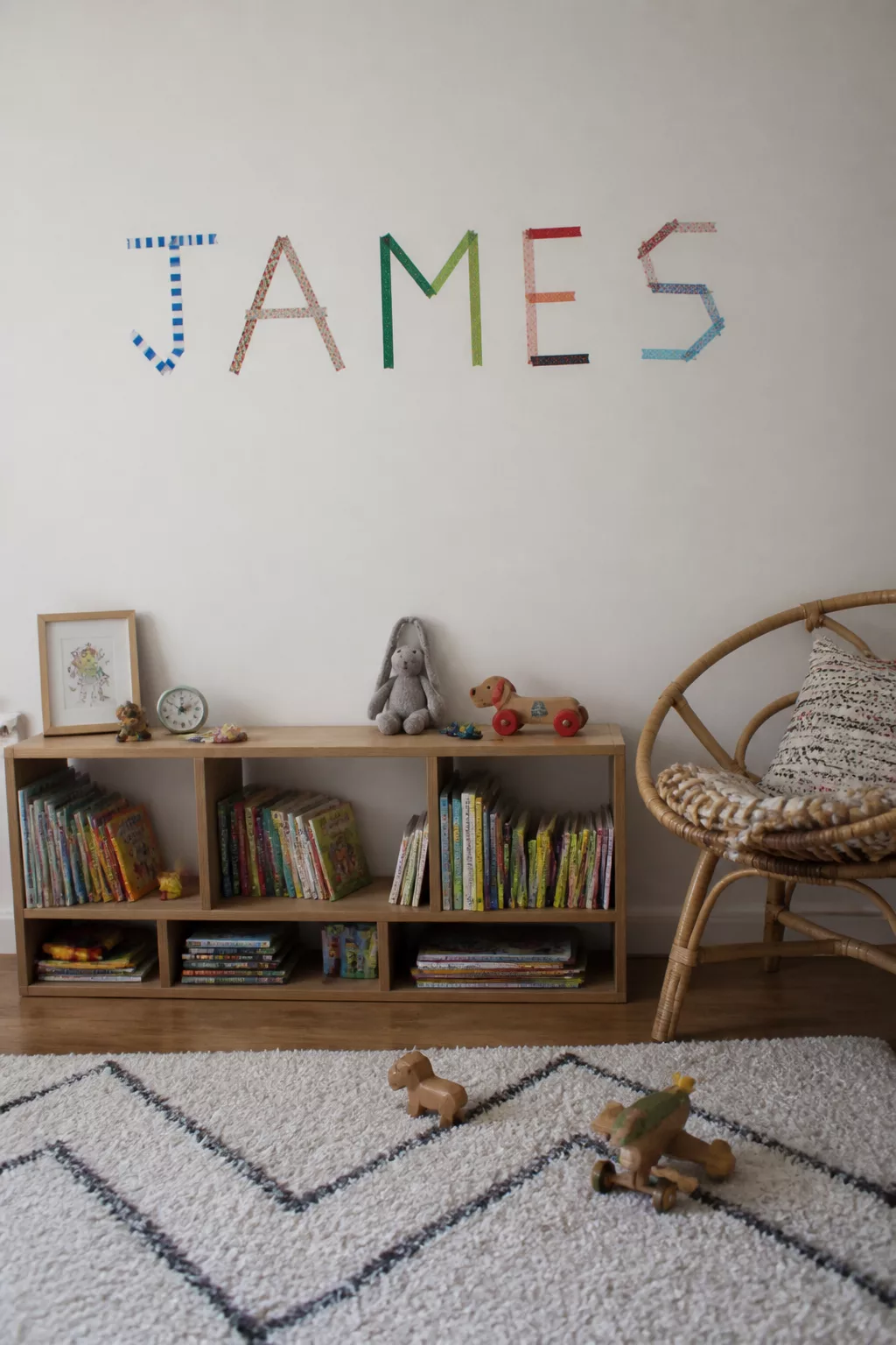 Colorful washi tape nursery name sign spelling JAMES above a wood bookshelf — easy DIY nursery name signs idea.
