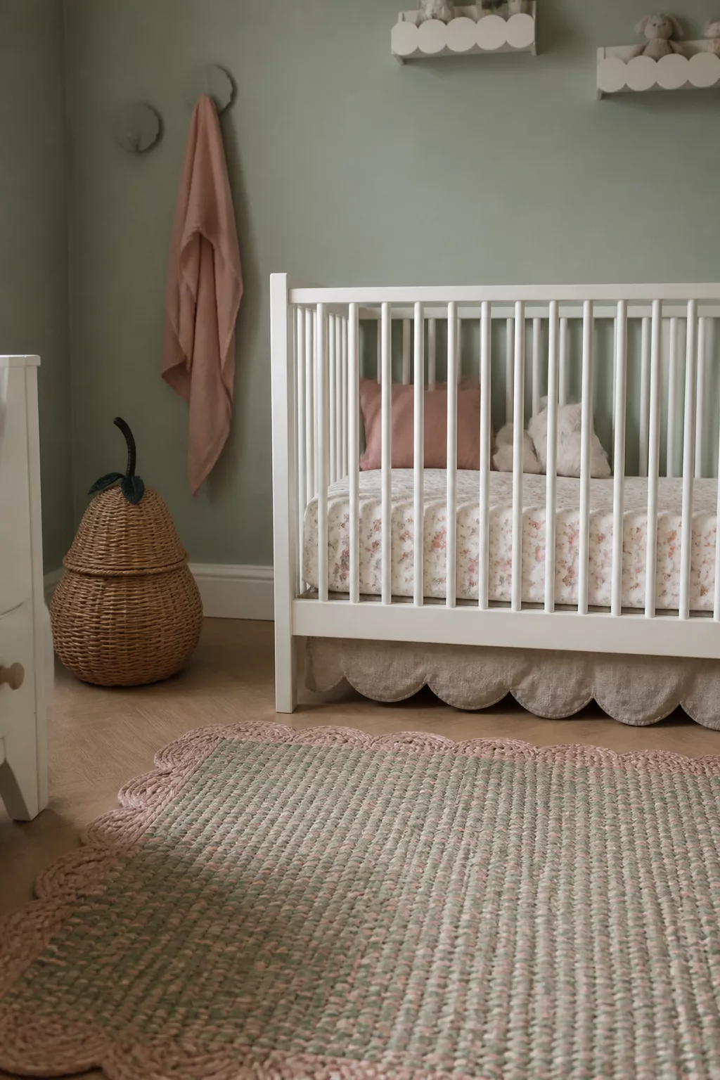 Sage green baby girl nursery ideas with scalloped crib skirt, blush pillows, and wicker pear basket