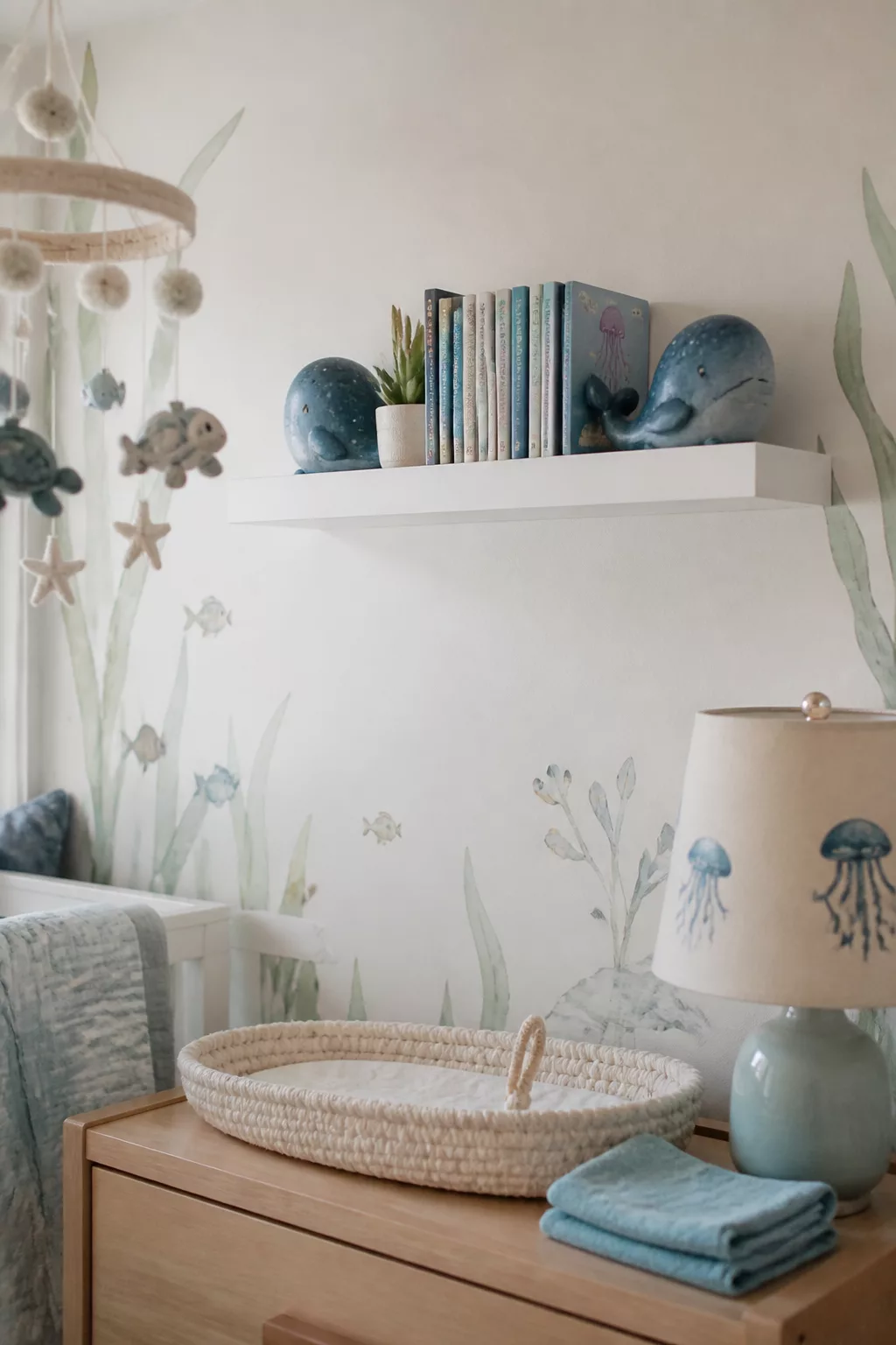 Whale bookends on a floating shelf above a dresser in an under the sea nursery with ocean mural wallpaper